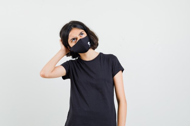 Kleines Mädchen, das im schwarzen T-Shirt, in der Maske und in der nachdenklichen Vorderansicht oben schaut.