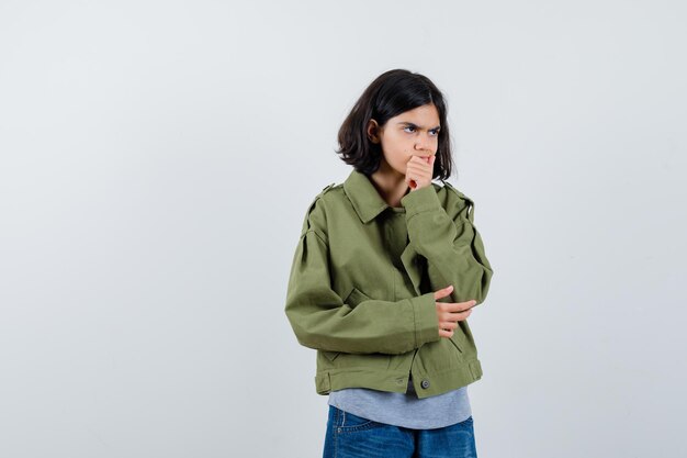 Kleines Mädchen, das in denkender Haltung in Mantel, T-Shirt, Jeans steht und nachdenklich aussieht. Vorderansicht.