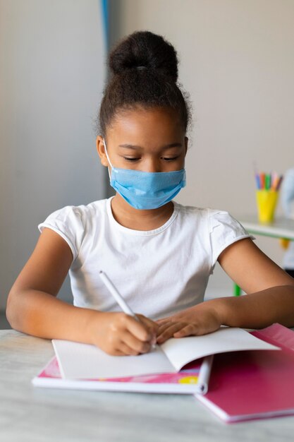 Kleines Mädchen, das in ein Notizbuch schreibt, während es eine medizinische Maske trägt