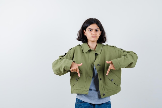 Kleines Mädchen, das in Mantel, T-Shirt, Jeans nach unten zeigt und selbstbewusst aussieht. Vorderansicht.