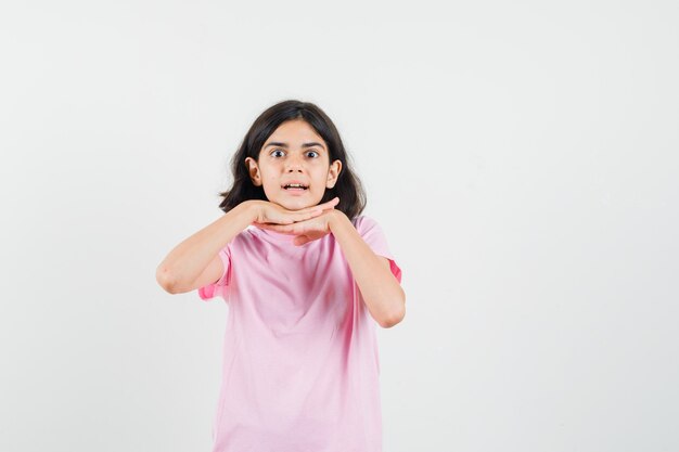 Kleines Mädchen, das Kinn auf Hände im rosa T-Shirt stützt und neugierig schaut. Vorderansicht.