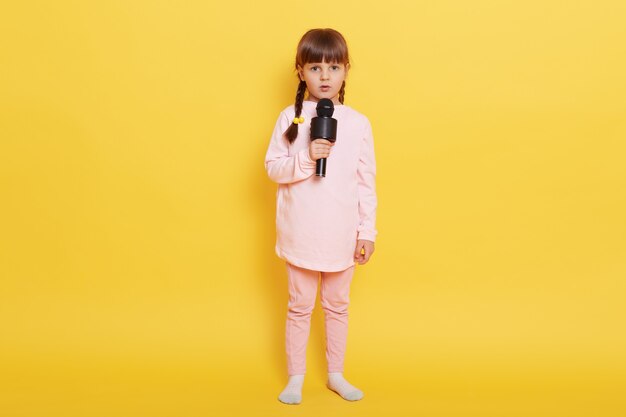 Kleines Mädchen, das Lied mit Kamera und ernstem Gesichtsausdruck singt, schaut Kamera mit besorgtem Blick an, verwirrt, um Leistung zu arrangieren, lässige Kleidung tragend, lokalisiert über gelbem Hintergrund.