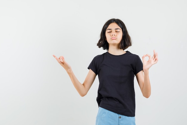 Kleines Mädchen, das Meditation mit geschlossenen Augen im schwarzen T-Shirt, in den Shorts tut und entspannt schaut. Vorderansicht.