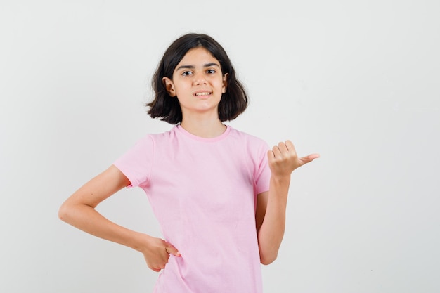 Kleines Mädchen, das mit Daumen im rosa T-Shirt beiseite zeigt und selbstbewusst, Vorderansicht schaut.