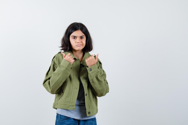 Kleines Mädchen, das mit Daumen in Mantel, T-Shirt, Jeans zur Seite zeigt und zögerlich aussieht. Vorderansicht.