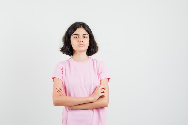 Kleines Mädchen, das mit verschränkten Armen im rosa T-Shirt steht und ernsthafte Vorderansicht sieht.