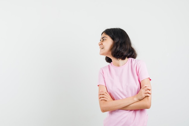 Kleines Mädchen, das mit verschränkten Armen im rosa T-Shirt steht und fröhlich, Vorderansicht schaut.
