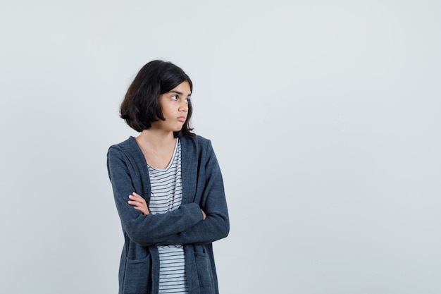 Kleines Mädchen, das mit verschränkten Armen im T-Shirt, in der Jacke steht und zögernd schaut