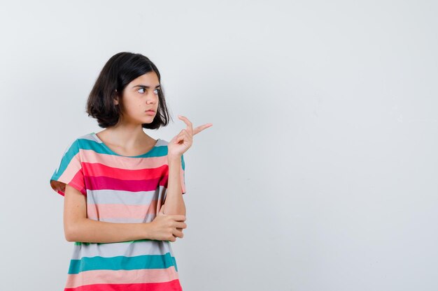 Kleines Mädchen, das nach rechts zeigt, während es die Hand am Ellbogen in T-Shirt, Jeans hält und fokussiert aussieht, Vorderansicht.