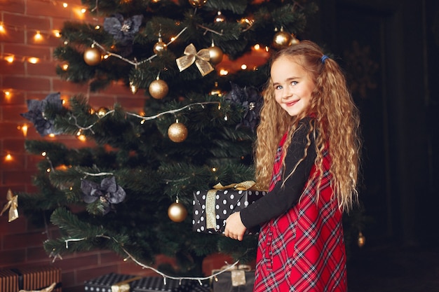 Kleines Mädchen, das nahe Weihnachtsbaum mit Geschenk steht