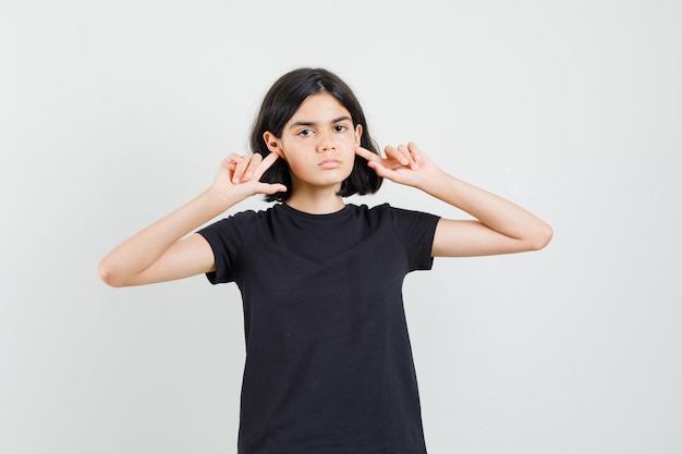 Kleines Mädchen, das Ohren mit den Fingern im schwarzen T-Shirt, in den kurzen Hosen und im ernsthaften Aussehen verstopft. Vorderansicht.