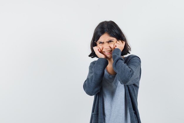 Kleines Mädchen, das Ohren mit den Fingern in T-Shirt, Jacke verstopft und gereizt aussieht,