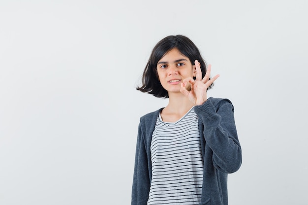 Kleines Mädchen, das ok Geste in T-Shirt, Jacke tut und selbstbewusst aussieht.