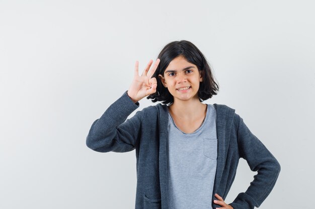 Kleines Mädchen, das ok Geste in T-Shirt, Jacke zeigt und selbstbewusst aussieht.