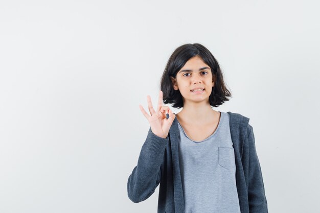 Kleines Mädchen, das ok Geste in T-Shirt, Jacke zeigt und selbstbewusst aussieht.