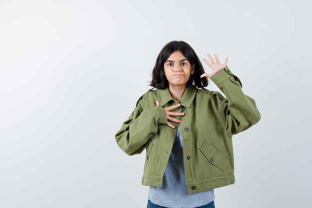 Kleines Mädchen, das Palme zeigt, während es die Hand auf der Brust in Mantel, T-Shirt, Jeans hält und selbstbewusst aussieht, Vorderansicht.