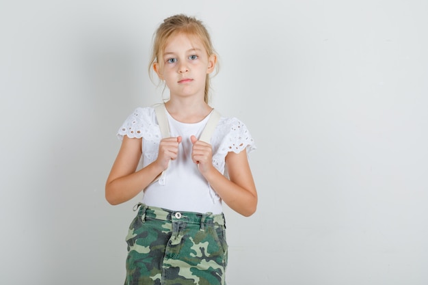 Kleines Mädchen, das Rucksack im weißen T-Shirt steht und trägt