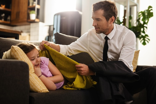Kleines Mädchen, das sich auf das Sofa legt, während ihr Vater sie mit einer Decke zudeckt, bevor sie zur Arbeit geht. Der Mensch steht im Mittelpunkt