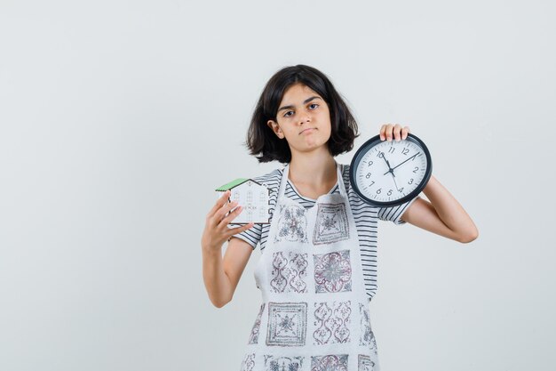 Kleines Mädchen, das Uhr und Hausmodell im T-Shirt, in der Schürze hält und nachdenklich schaut,