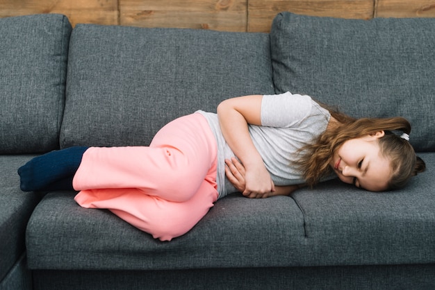 Kleines Mädchen, das unter den Magenschmerzen leiden auf grauem Sofa leidet