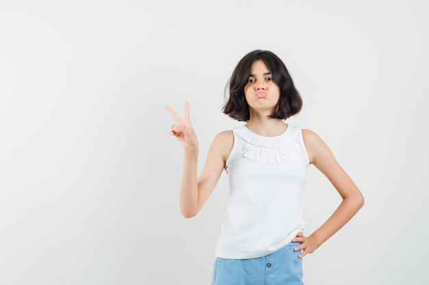 Kleines Mädchen, das V-Zeichen zeigt, schmollende Lippen in weißer Bluse, Shorts und selbstbewusst aussehend, Vorderansicht.