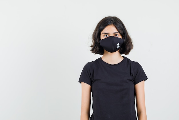 Kleines Mädchen, das vorne im schwarzen T-Shirt, in der Maske und in der ruhigen Vorderansicht schaut.