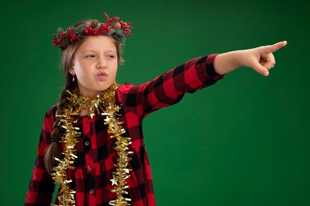 Kleines Mädchen, das Weihnachtskranz im karierten Kleid mit Lametta um den Hals trägt und beiseite schaut mit ernstem Gesicht, das mit Zeigefinger auf etwas zeigt