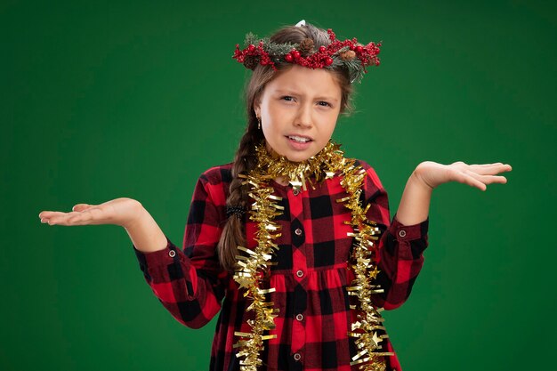 Kleines Mädchen, das Weihnachtskranz im karierten Kleid mit Lametta um den Hals trägt verwirrt verwirrte Arme zu den Seiten