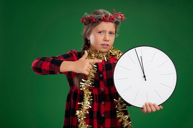 Kleines Mädchen, das Weihnachtskranz in kariertem Kleid mit Lametta um den Hals trägt und eine Wanduhr hält, die mit dem Zeigefinger darauf zeigt, dass sie verwirrt über der grünen Wand steht?