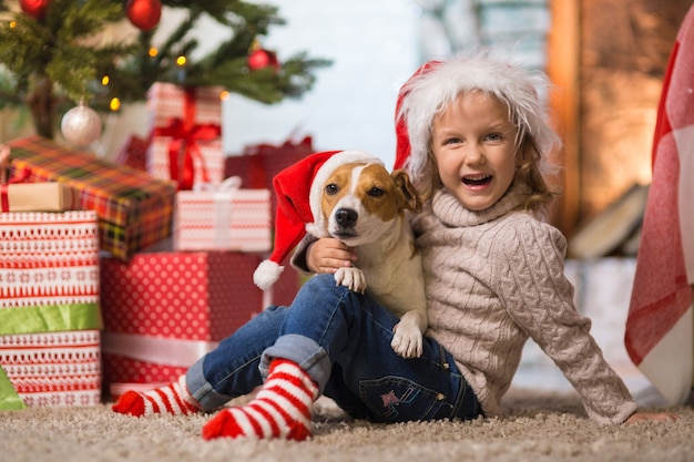 Kleines mädchen, das zu hause mit einem hund jack russell ein frohes weihnachtsfest am kamin feiert