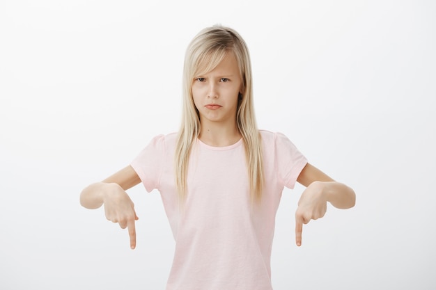 Kostenloses Foto kleines mädchen hasst abwasch. porträt einer missfallenen angewiderten süßen kleinen tochter mit blonden haaren, die nach unten zeigt und die stirn runzelt, beleidigt ist, abneigung und ärger über die graue wand ausdrückt