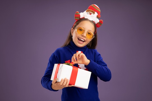 Kleines Mädchen im blauen Rollkragenpullover mit roter Krawatte und lustigem Weihnachtsrand auf dem Kopf, der ein Geschenk mit einem Lächeln im Gesicht hält, das glücklich und fröhlich über der lila Wand steht