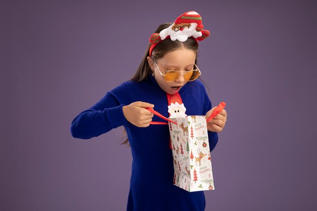 Kleines Mädchen im blauen Rollkragenpullover mit roter Krawatte und lustigem Weihnachtsrand auf Kopf, der Papiertüte mit Weihnachtsgeschenk hält, das innen schaut, das über lila Hintergrund steht