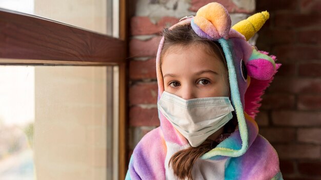 Kleines Mädchen im Dinosaurieranzug zu Hause mit Gesichtsmaske während der Quarantäne