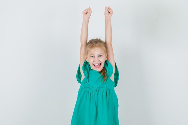 Kleines Mädchen im grünen Kleid, das Arme hebt und schreit und glücklich aussieht