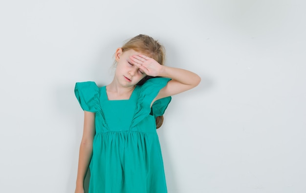 Kleines Mädchen im grünen Kleid, das Hand auf Stirn hält und schläfrig aussieht