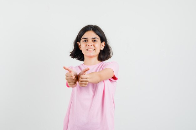 Kleines Mädchen im rosa T-Shirt, das doppelte Daumen oben zeigt und fröhlich, Vorderansicht schaut.