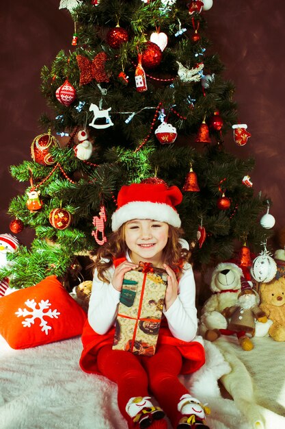 Kleines Mädchen im roten Kleid sitzt mit einem Präsentkarton vor einem Weihnachtsbaum