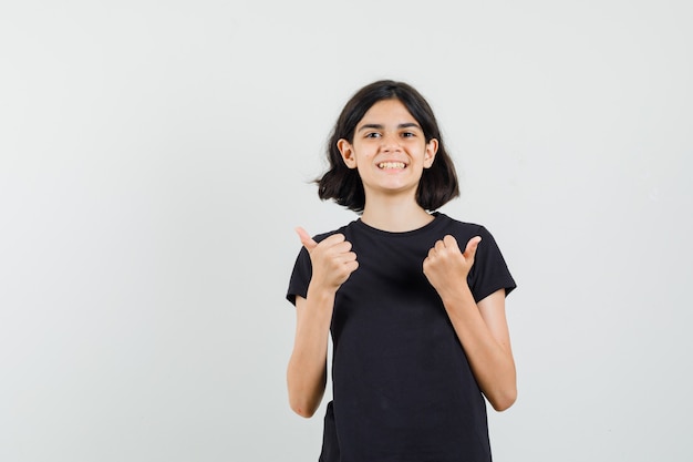 Kleines Mädchen im schwarzen T-Shirt, das doppelte Daumen oben zeigt und glücklich schaut, Vorderansicht.