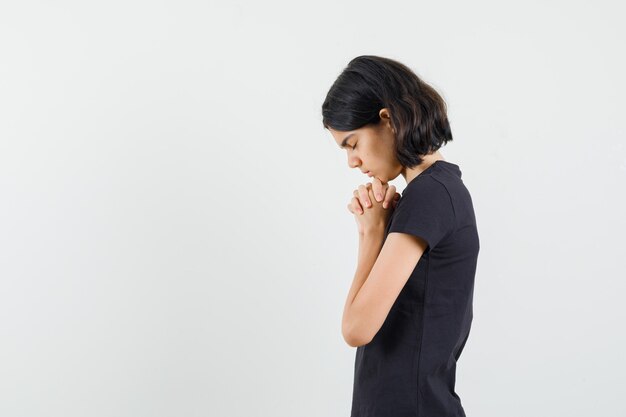 Kleines Mädchen im schwarzen T-Shirt, das Hände in der Gebetsgeste fasst und hoffnungsvoll aussieht.
