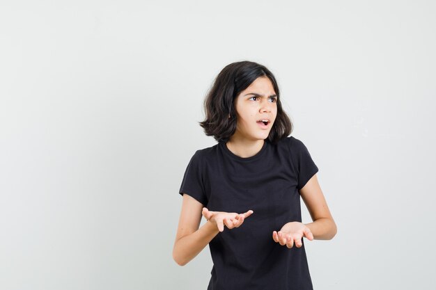 Kleines Mädchen im schwarzen T-Shirt, das Hände in verwirrter Geste, Vorderansicht hält.