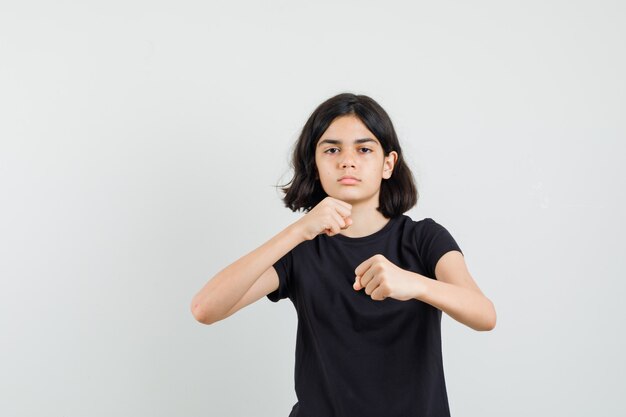 Kleines Mädchen im schwarzen T-Shirt, das in der Boxerhaltung steht und strenge Vorderansicht schaut.