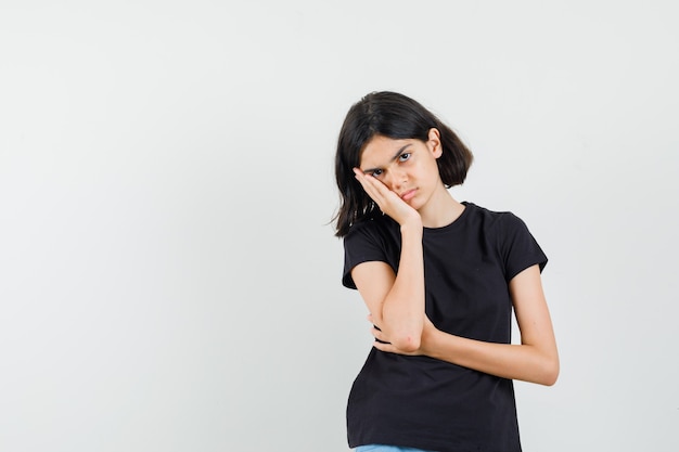 Kleines Mädchen im schwarzen T-Shirt, das Wange auf erhabener Handfläche lehnt und traurige Vorderansicht schaut.