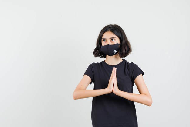 Kleines Mädchen im schwarzen T-Shirt, Maske, die namaste Geste zeigt, Vorderansicht.