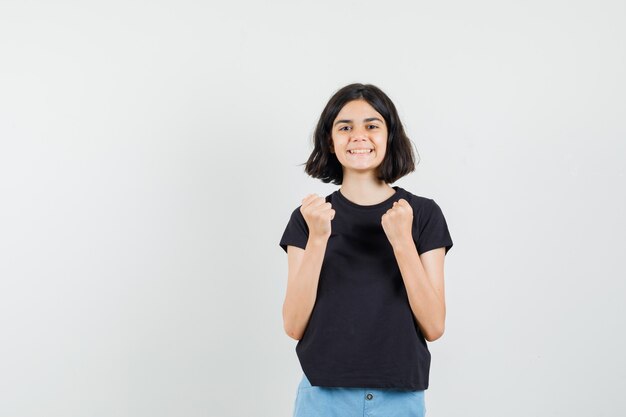 Kleines Mädchen im schwarzen T-Shirt, Shorts, die Siegergeste zeigen und fröhlich schauen, Vorderansicht.