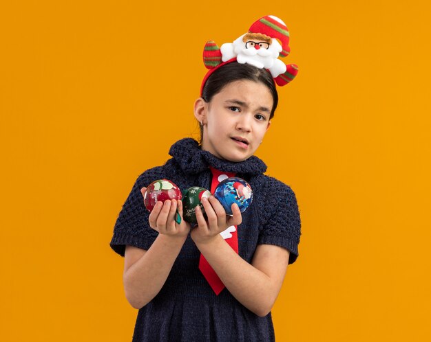 Kleines Mädchen im Strickkleid, das rote Krawatte mit lustigem Rand auf Kopf hält, der Weihnachtskugeln hält, die besorgt schauen