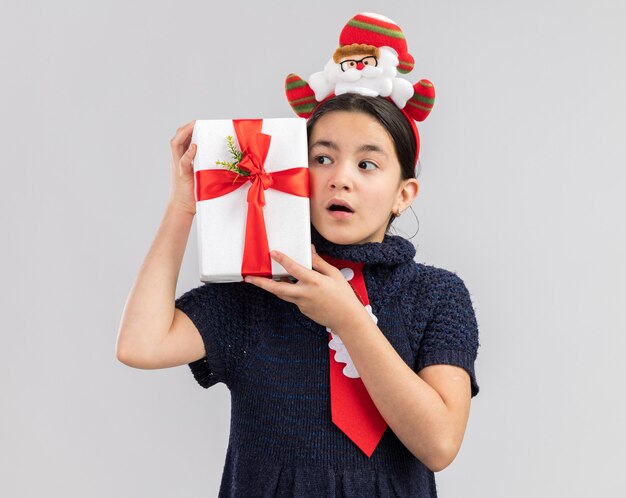 Kleines Mädchen im Strickkleid, das rote Krawatte mit lustigem Weihnachtsrand auf Kopf hält, der Weihnachtsgeschenk hält, das beiseite überrascht schaut