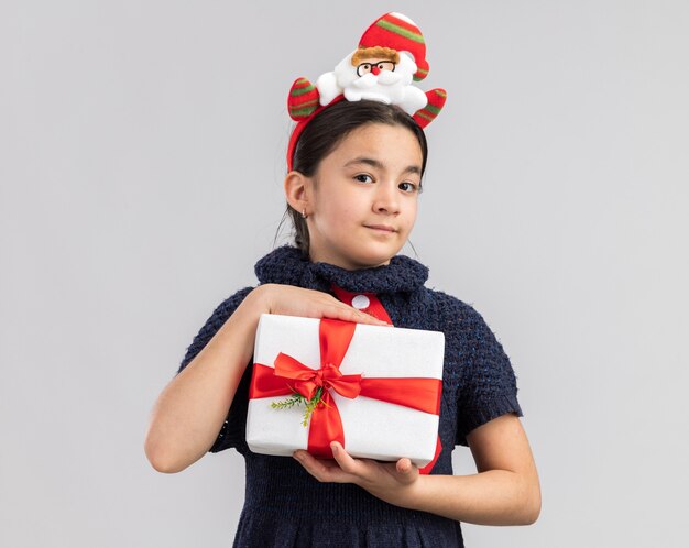 Kleines Mädchen im Strickkleid, das rote Krawatte mit lustigem Weihnachtsrand auf Kopf hält, der Weihnachtsgeschenk hält, das zuversichtlich lächelt