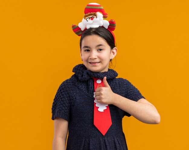 Kleines Mädchen im Strickkleid mit roter Krawatte mit lustigem Weihnachtsrand auf dem Kopf, der glücklich und psitiv zeigt Daumen zeigt