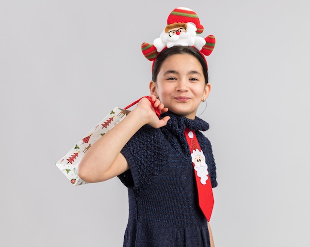 Kleines Mädchen im Strickkleid trägt rote Krawatte mit lustigem Weihnachtsrand auf Kopf, der Papiertüte mit Weihnachtsgeschenk hält, das glücklich und positiv schaut